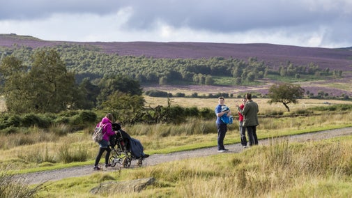 Three people stood talking in a group with two other people travelling towards them. One of the people is walking and the other is in a wheelchair. There are views of the rural landscape including patches of purple heather on the moorland in the distance.